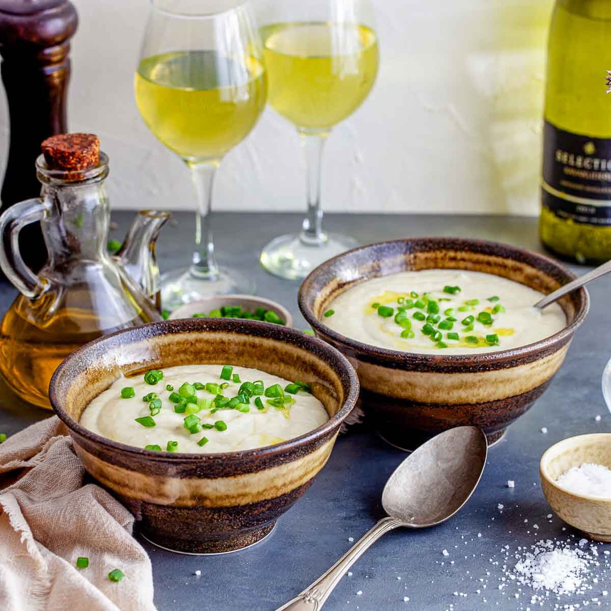 Two bowls of creamy potato-leek soup (vichyssoise), garnished with chopped chives and a drizzle of oil, served with wine in the background, ready to enjoy