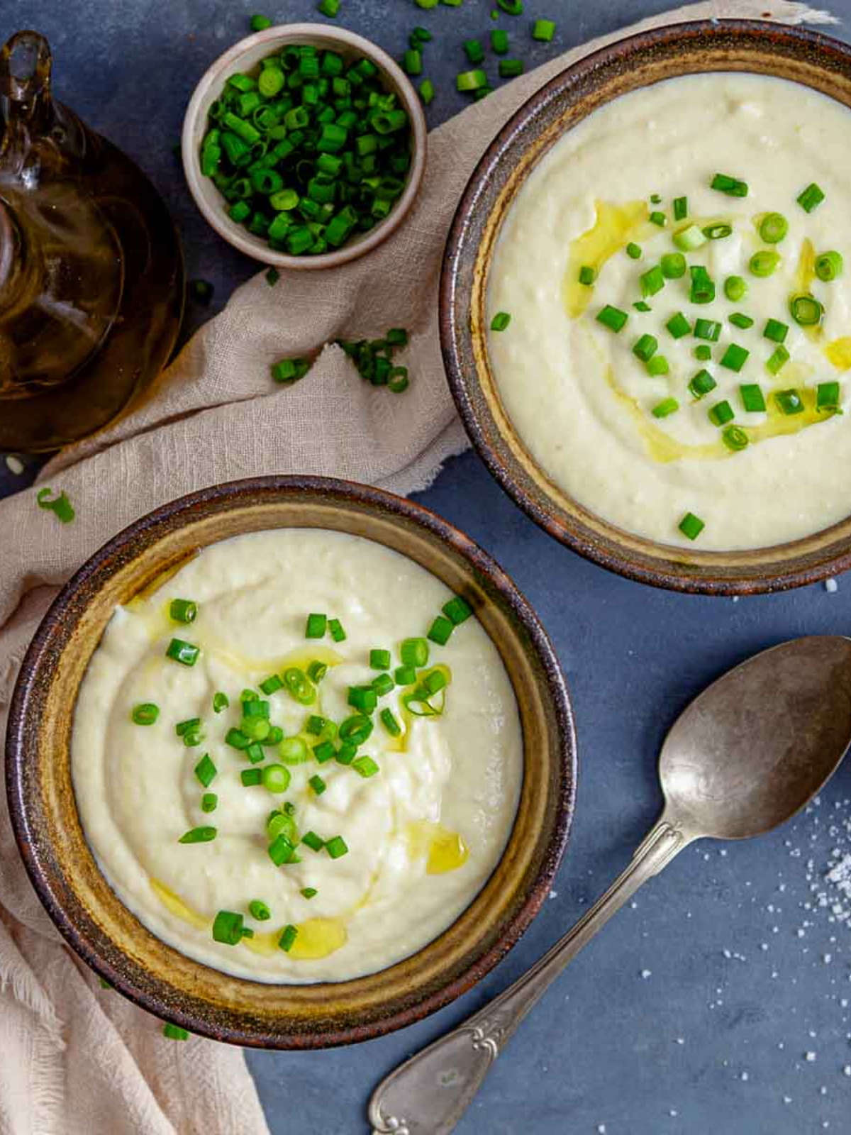 Two bowls of creamy soup topped with chopped green onions and a drizzle of olive oil, served with salt.