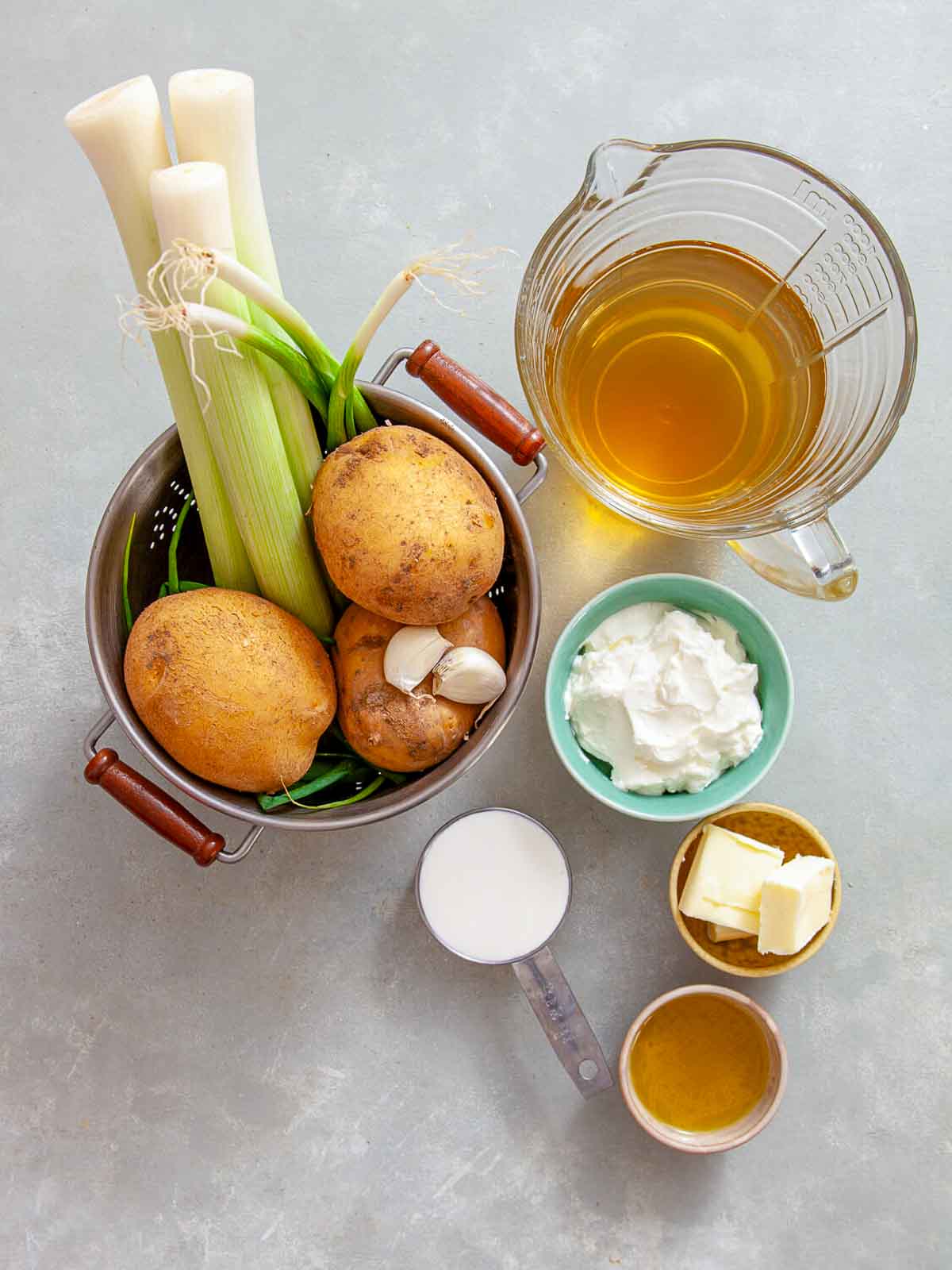 Ingredients for a creamy soup laid out on a surface, including leeks, potatoes, garlic, broth, cream, butter, and oil