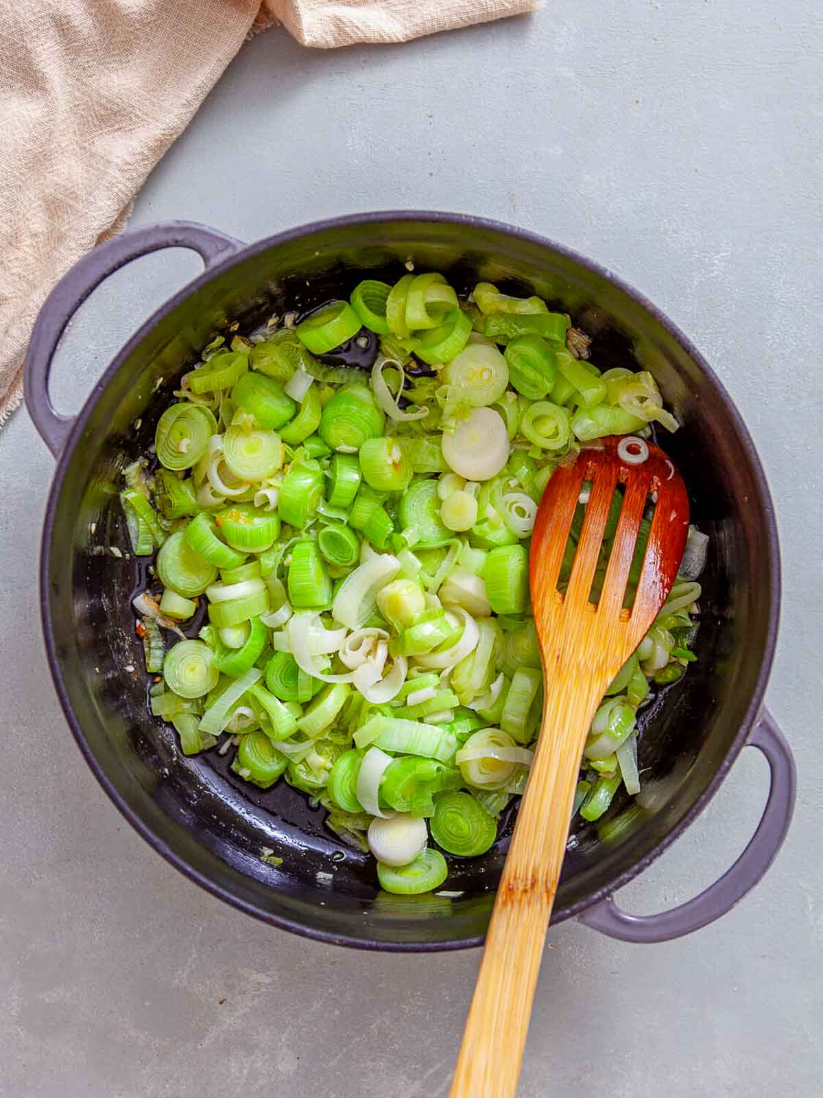 Sliced leeks being sautéed in a pot with oil, stirred with a wooden spoon