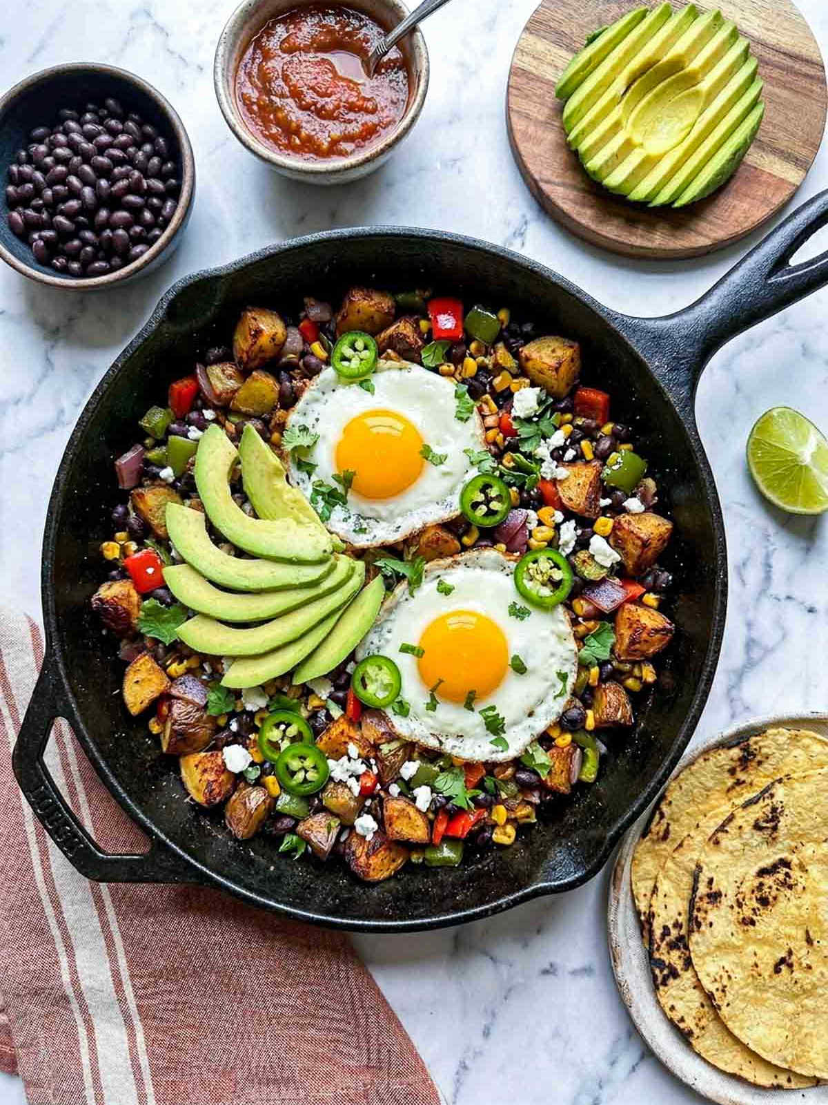 Overhead shot of a cast iron skillet with Mexican black bean breakfast hash — crispy diced potatoes, black beans, red and green bell pepper, red onion, and charred corn, topped with two sunny-side-up fried eggs, sliced avocado, crumbled cotija, sliced jalapeño, and fresh cilantro, on a white marble surface with a muted striped linen napkin, bowls of black beans and salsa, sliced avocado on a wooden board, charred corn tortillas, and halved lime.