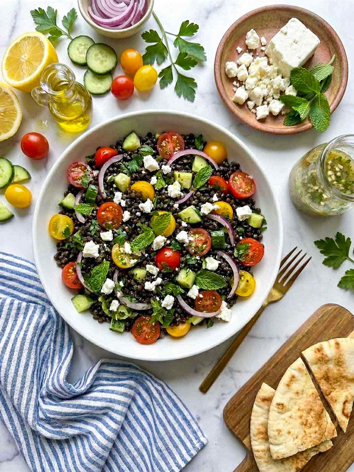 Mediterranean black lentil salad topped with feta, cucumber, tomatoes, red onion, parsley, and mint, surrounded by fresh ingredients and pita bread on a marble surface.