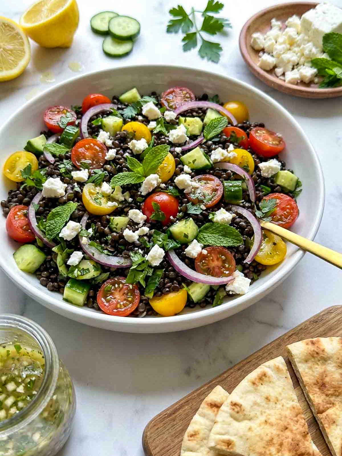 Close-up view of black lentil cold salad with cucumber, cherry tomatoes, red onion, feta cheese, and fresh herbs in a large serving bowl.