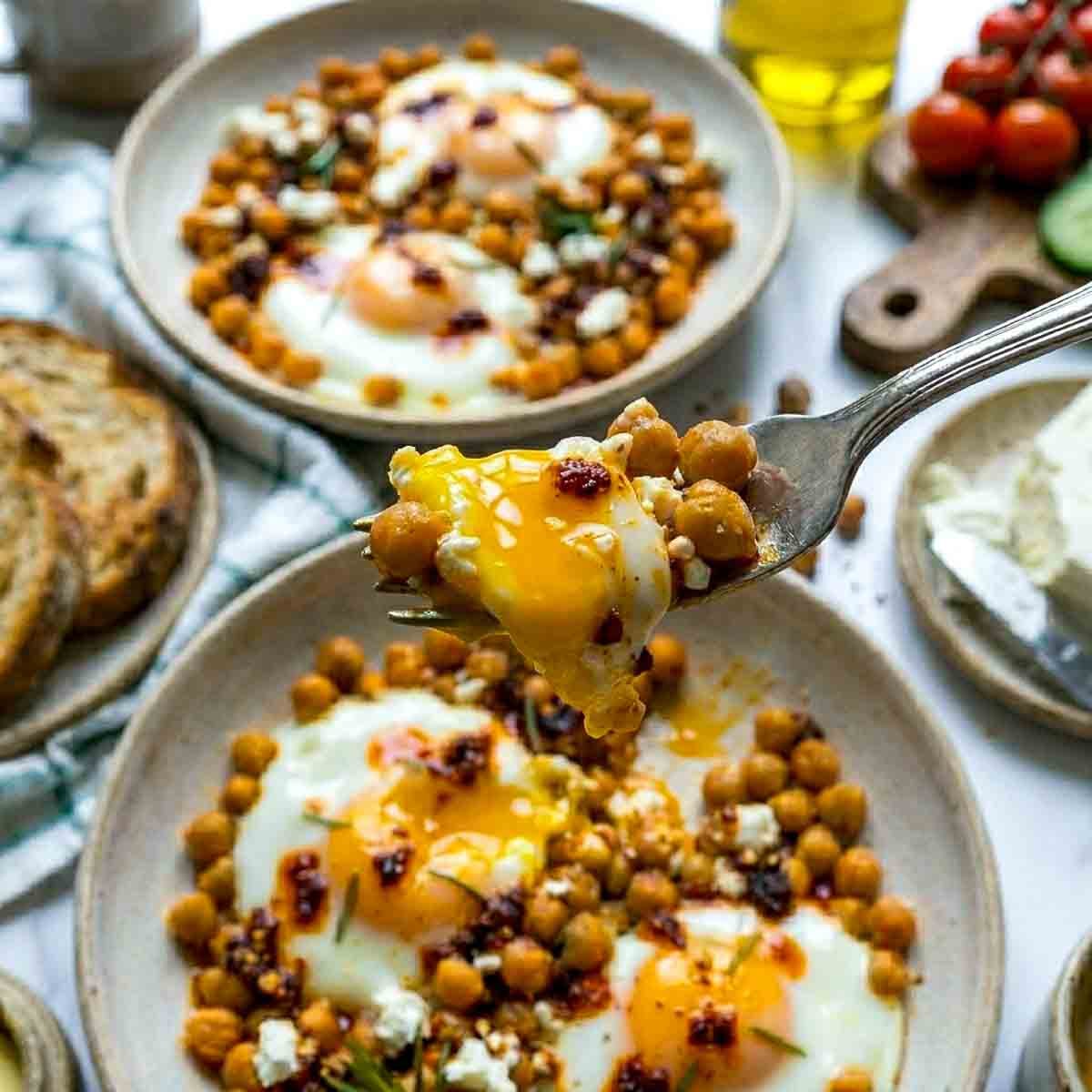 A fork lifting a bite of chickpea fried eggs with a runny yolk over a bowl of crispy chickpeas and eggs topped with chili oil and crumbled feta, with a second serving bowl in the background.