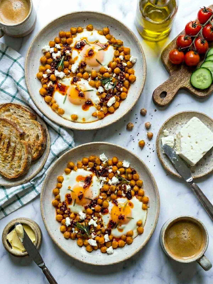 Overhead flat lay of two bowls of chickpea fried eggs with runny yolks, crumbled feta, chili oil, and fresh rosemary, served alongside toast, butter, coffee, cherry tomatoes, and sliced cucumber on a marble surface.