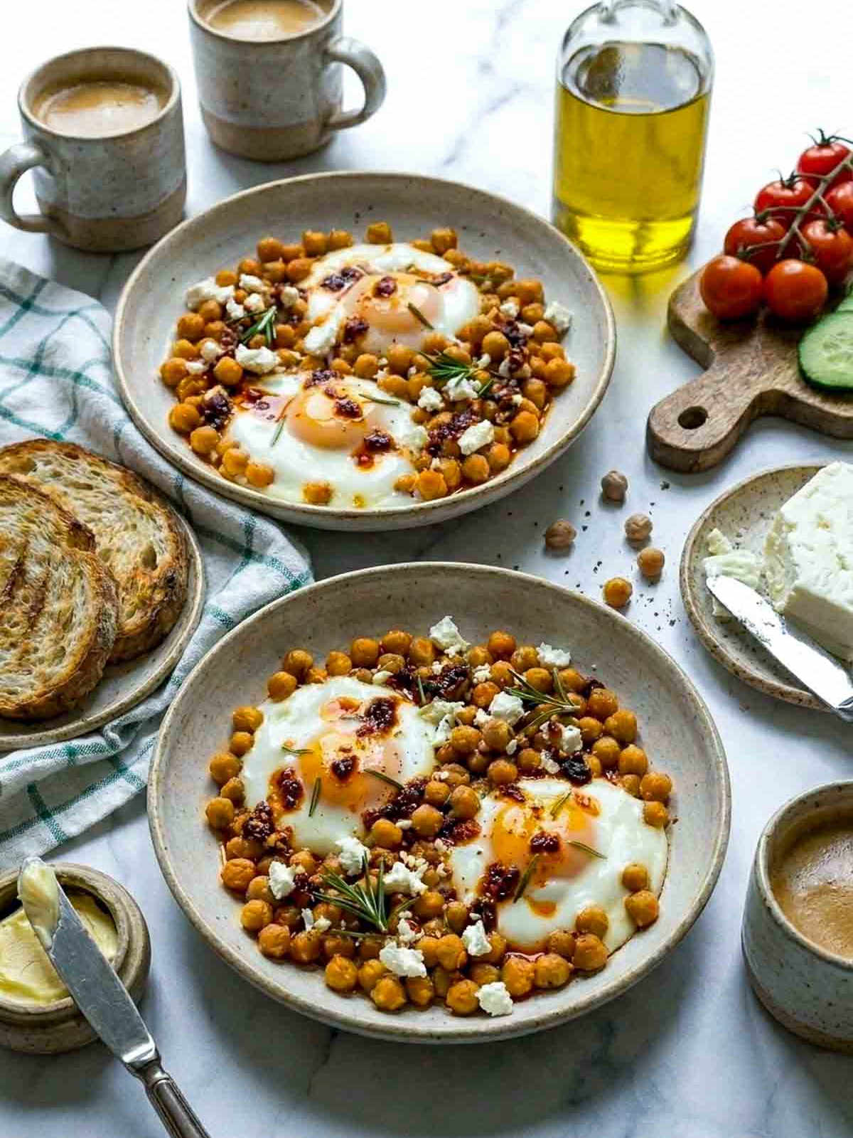 Two rustic speckled bowls of chickpea fried eggs topped with feta and chili oil on a marble surface, surrounded by toasted bread, coffee mugs, olive oil, and cherry tomatoes on a wooden board.