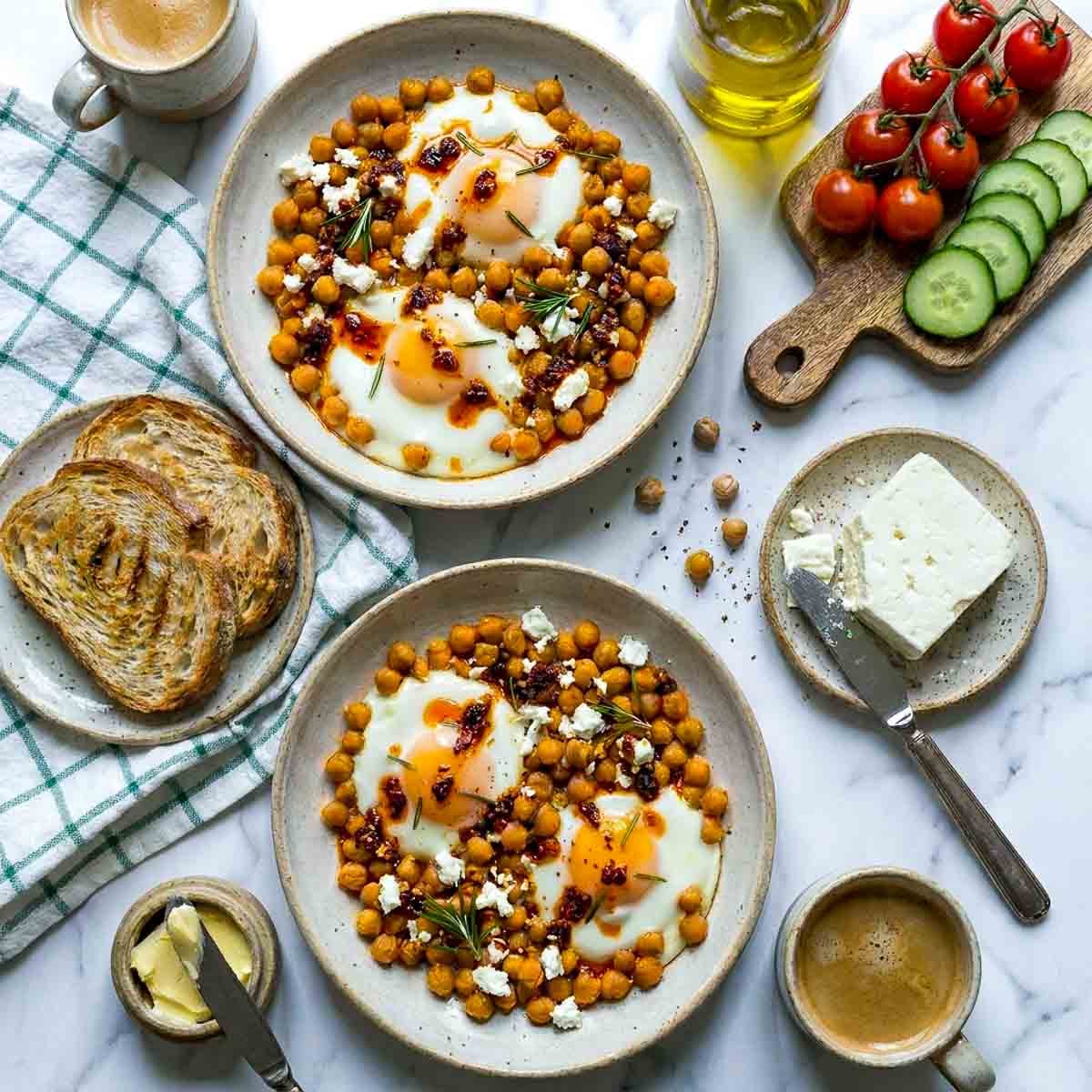 Top-down view of two bowls of chickpea fried eggs with runny yolks, crumbled feta, rosemary, and chili oil drizzle, styled with toast, coffee, butter, and fresh cherry tomatoes on a marble background.