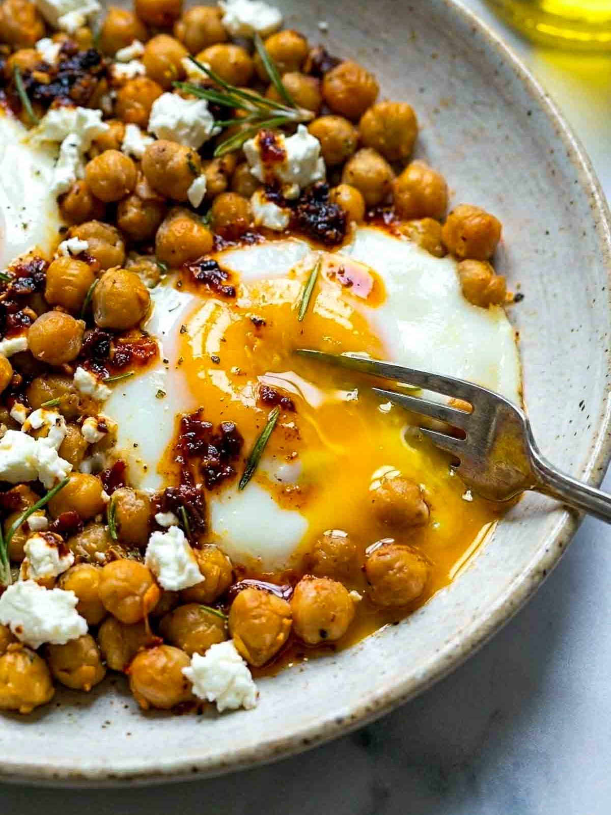 Close-up of a speckled bowl of chickpea fried eggs with a broken runny yolk, crispy golden chickpeas, crumbled feta, chili oil, and fresh rosemary, with a fork resting in the bowl.