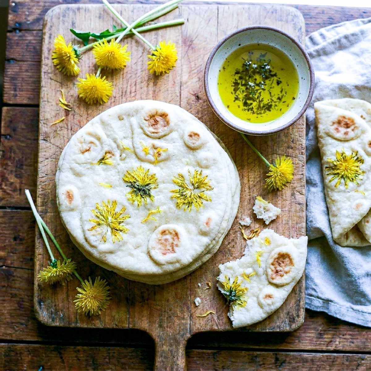 Overhead view of a stack of soft round flatbreads with dandelion flowers pressed into the tops on a wooden board, a torn piece in the foreground showing a soft fluffy interior, a small bowl of herbed olive oil for dipping, fresh dandelion flowers scattered around, linen napkin, warm natural light
