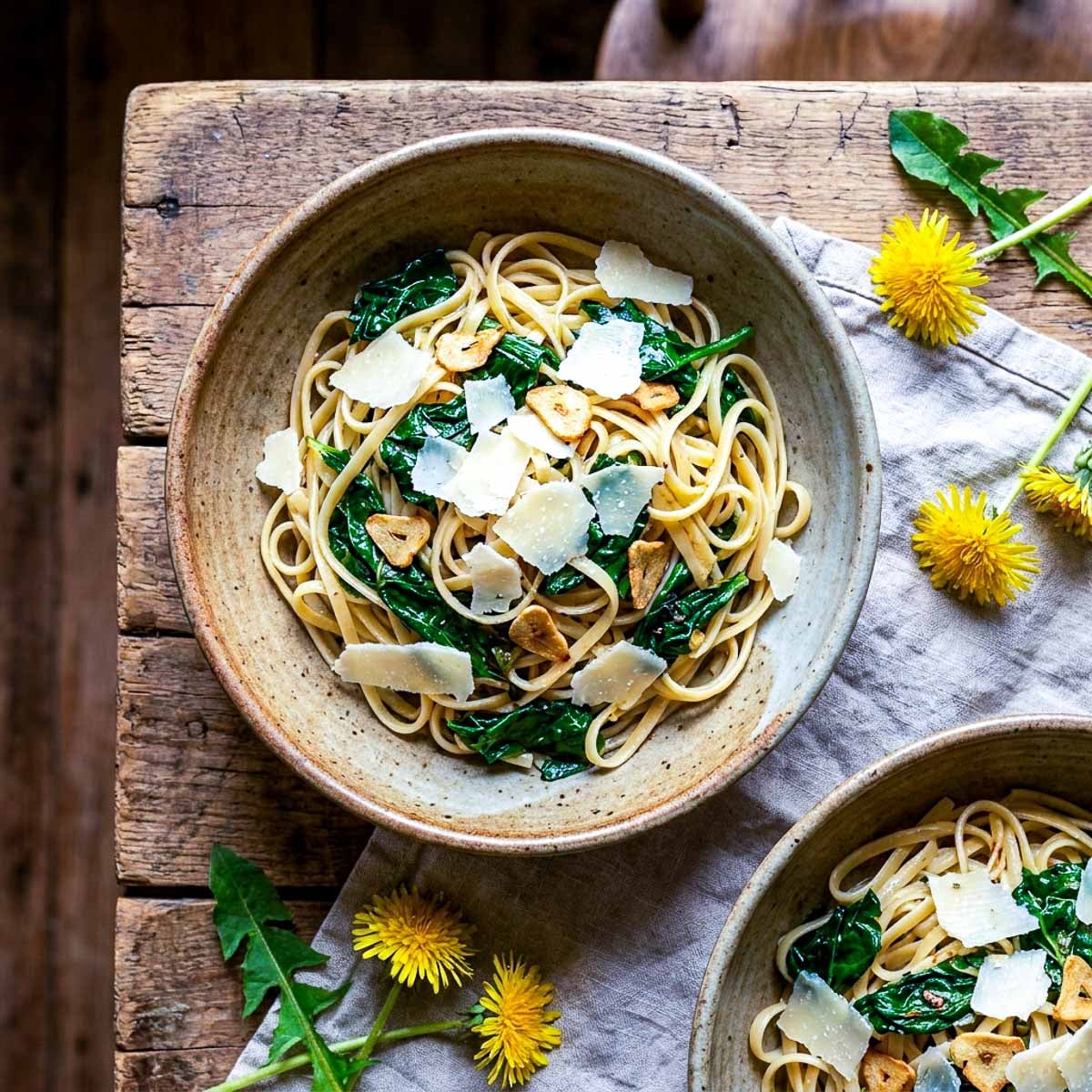 Overhead view of two rustic ceramic bowls of linguine tossed with wilted dandelion greens, golden garlic slivers, and shaved parmesan, fresh dandelion flowers and leaves scattered on a worn wooden surface, linen napkin