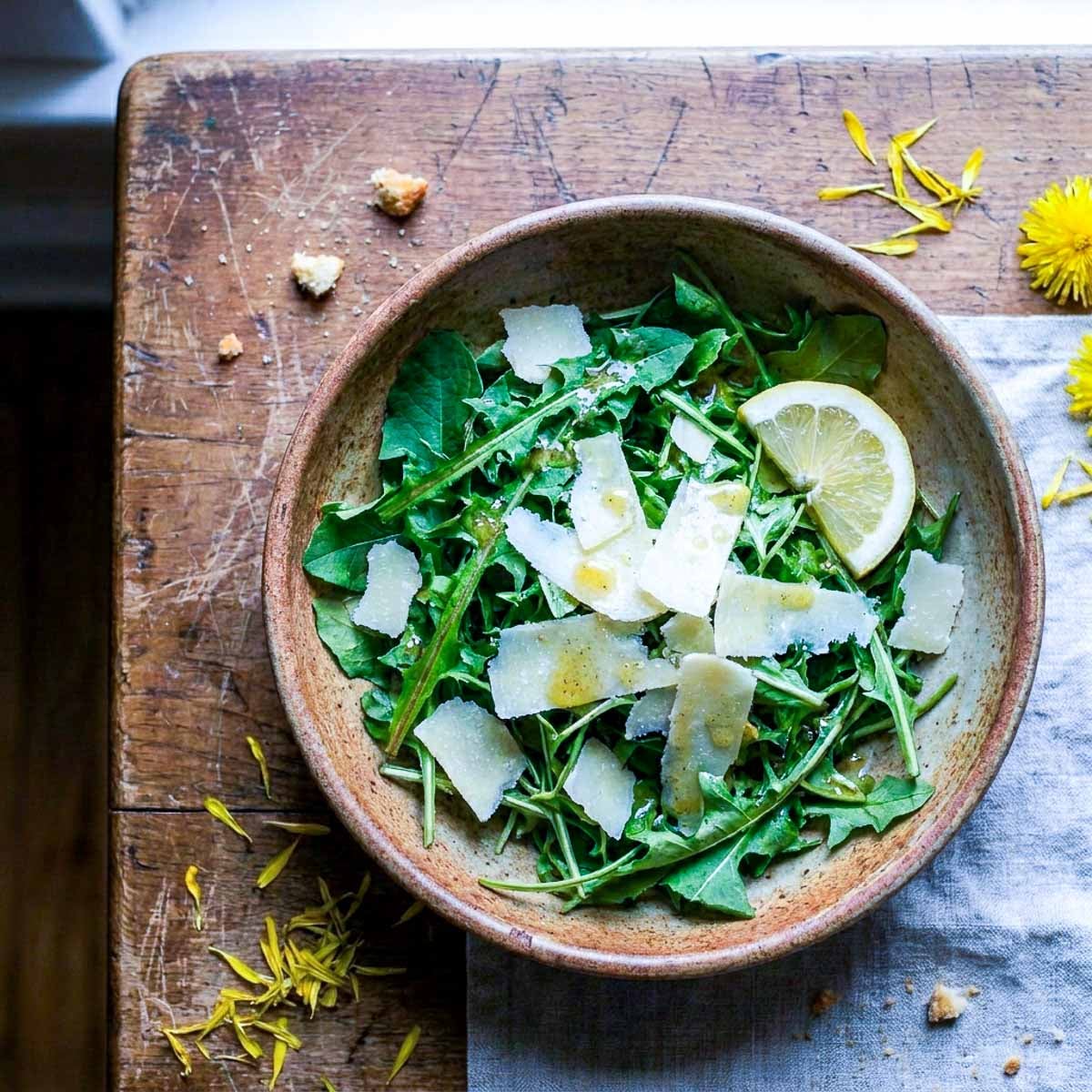 Overhead view of a rustic ceramic bowl filled with fresh dandelion greens, shaved parmesan, and a lemon wedge, scattered dandelion petals on a worn wooden surface, soft natural window light