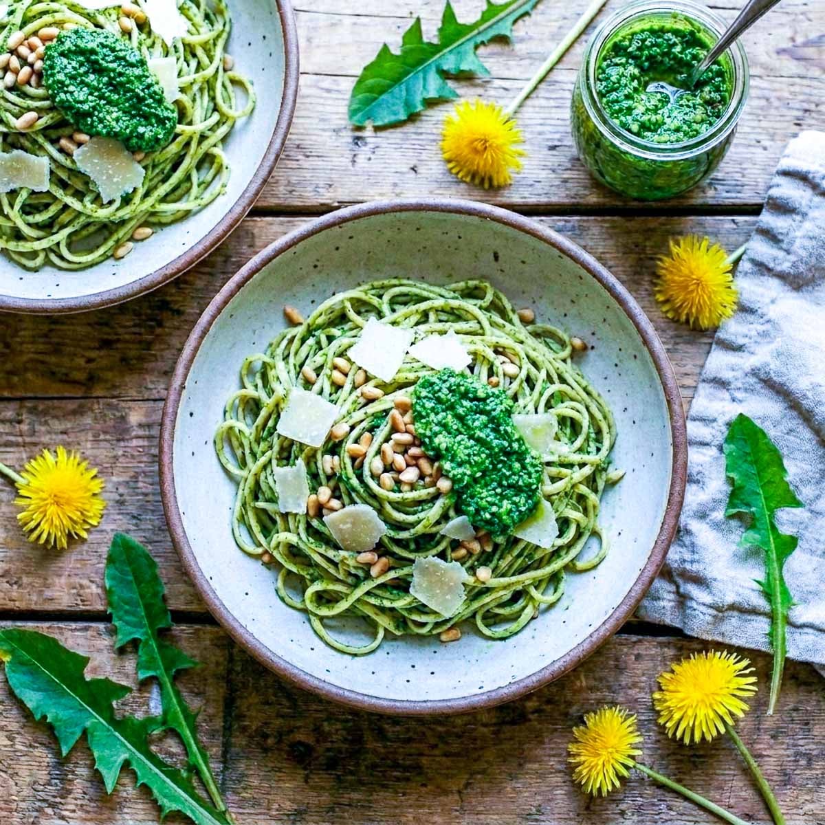 Overhead view of two rustic ceramic bowls of spaghetti coated in vibrant green dandelion pesto, pine nuts and shaved parmesan on top, a jar of pesto and fresh dandelion flowers and leaves scattered on a worn wooden surface, linen napkin