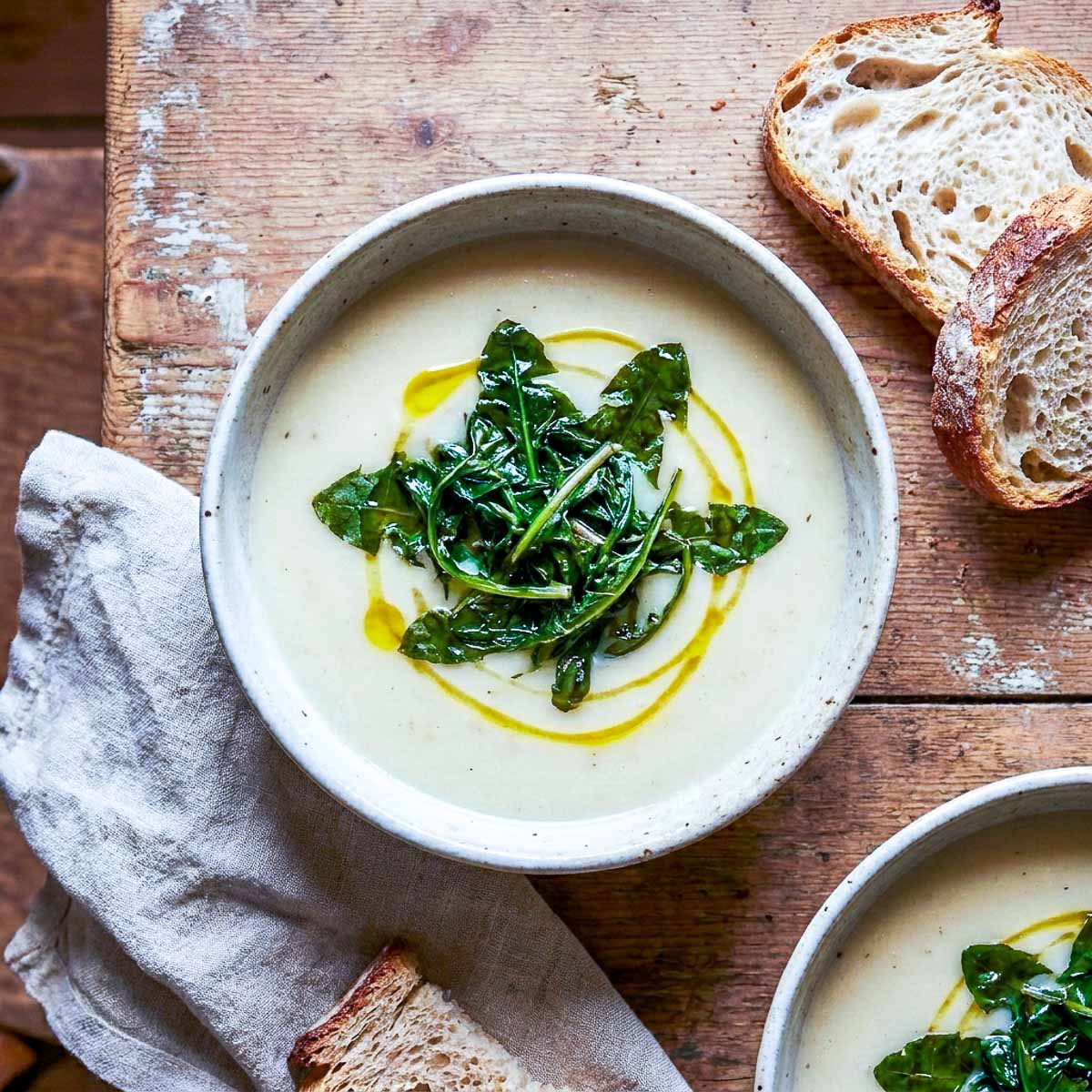 Overhead view of two rustic ceramic bowls of creamy potato soup topped with wilted dandelion greens and an olive oil drizzle, crusty bread slices and a linen napkin on a worn wooden surface