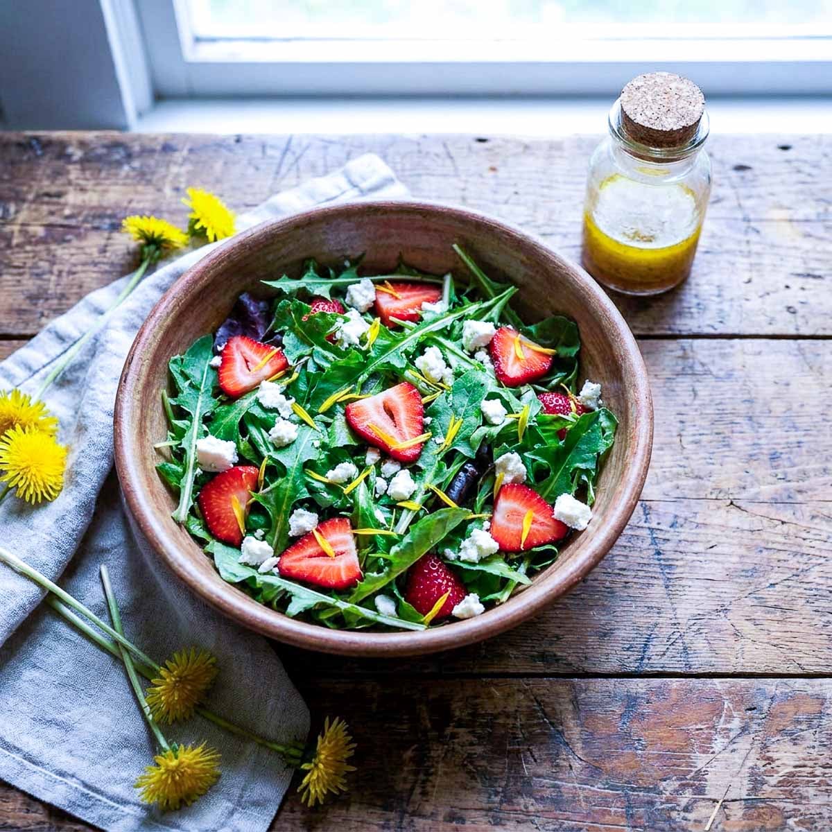 A rustic wooden bowl of dandelion greens with sliced strawberries, crumbled feta, and scattered dandelion petals, a small jar of golden dressing beside it, worn wooden table, soft spring window light
