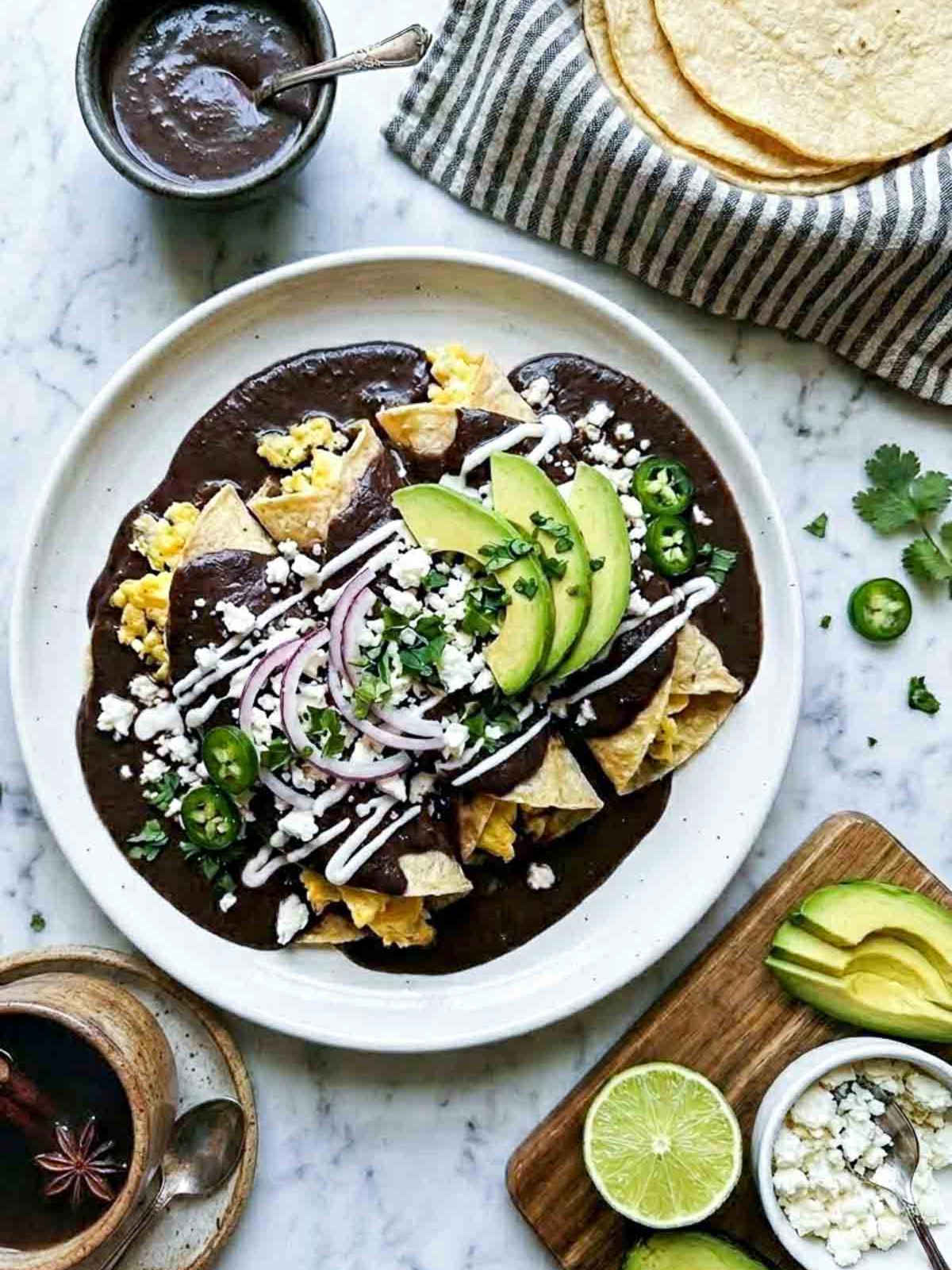 Overhead shot of a white ceramic plate with enfrijoladas — corn tortillas coated in a rich black bean sauce, filled with scrambled eggs, topped with crumbled cotija, sliced avocado, red onion rings, sliced jalapeño, crema drizzle, and fresh cilantro, on a white marble surface with a muted striped linen napkin, bowl of black bean sauce, stack of corn tortillas, sliced avocado and cotija on a wooden board, and halved lime.