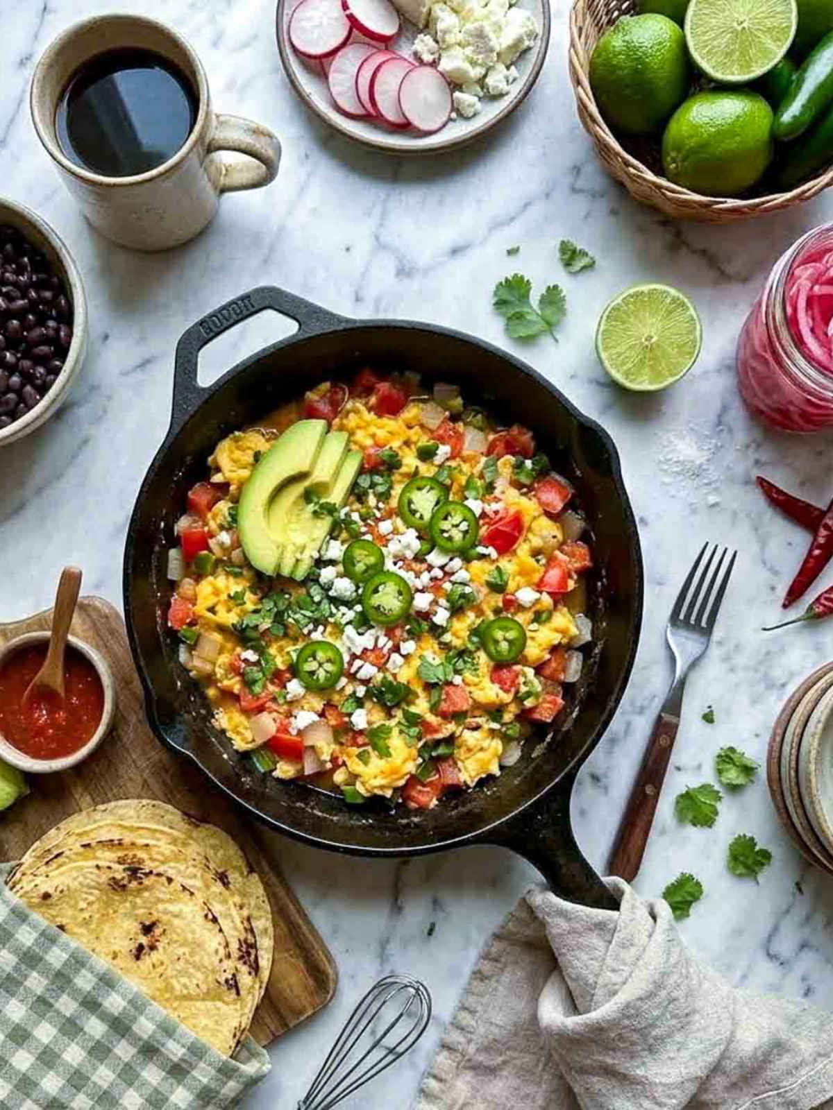 Overhead shot of a cast iron skillet with huevos a la mexicana — scrambled eggs cooked with diced tomato, white onion, and serrano pepper, topped with sliced avocado, crumbled cotija, sliced jalapeño, and fresh cilantro, on a white marble surface with a green check linen napkin, corn tortillas on a wooden board, bowl of salsa, black beans, coffee, pickled onions, dried chiles, and fresh limes in the background.