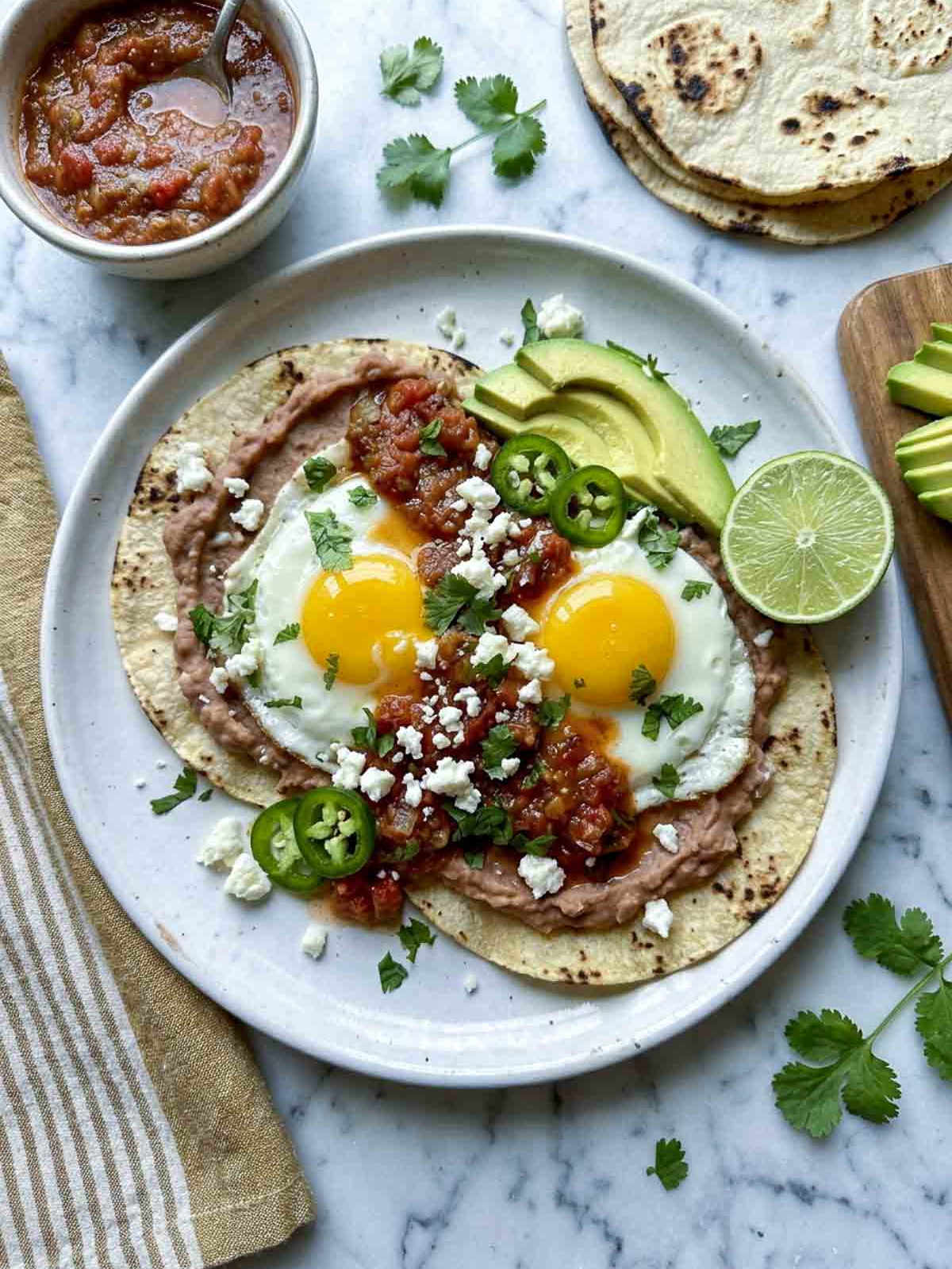Overhead shot of a white ceramic plate with huevos rancheros — two fried eggs on charred corn tortillas spread with refried beans, topped with chunky salsa roja, crumbled cotija, sliced jalapeño, avocado, and fresh cilantro, on a white marble surface with a muted gold striped linen napkin, bowl of salsa, stack of tortillas, and avocado slices in the background.