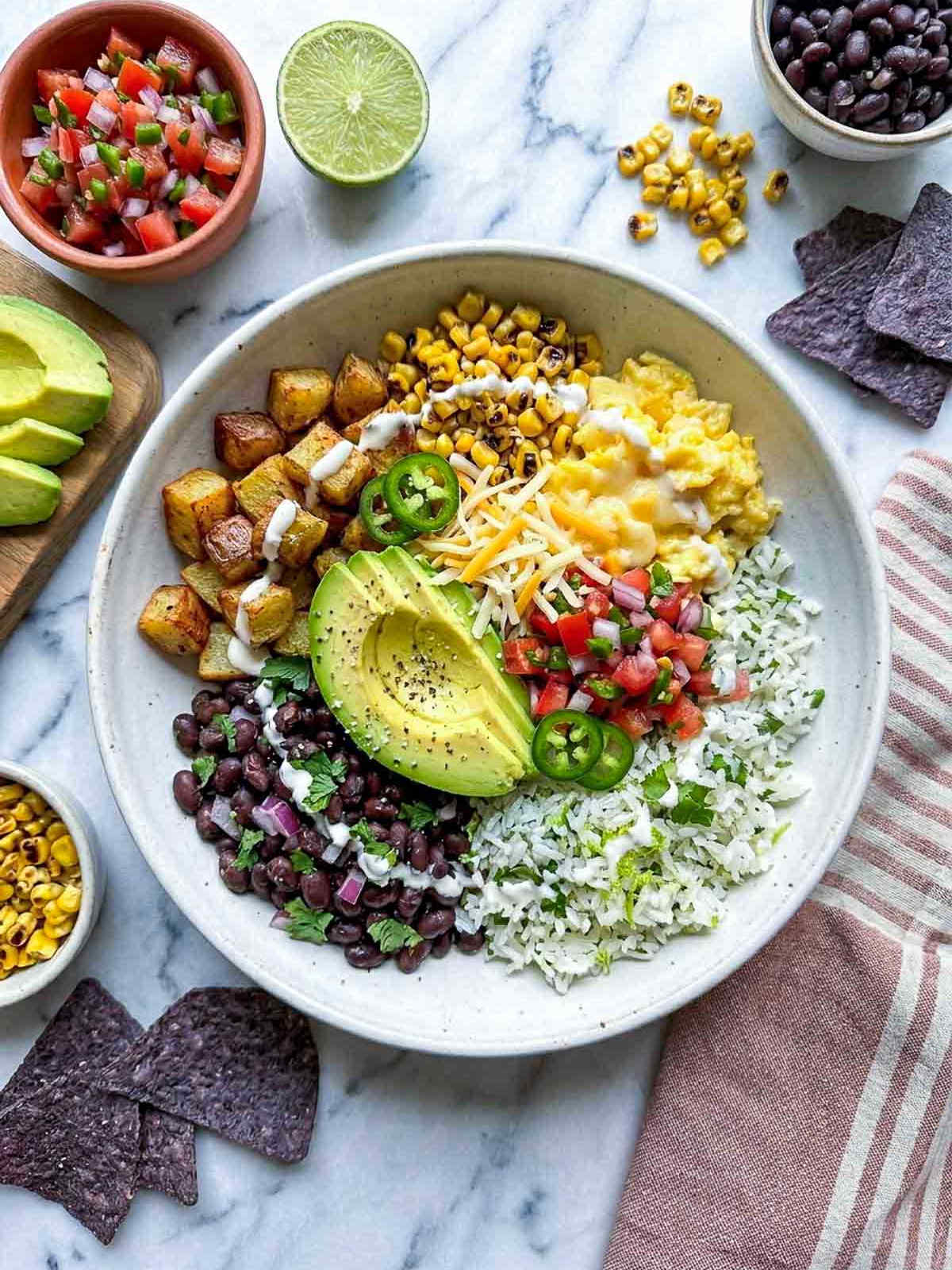 Overhead shot of a wide white ceramic bowl with a Mexican breakfast bowl — cilantro lime rice, black beans, scrambled eggs, roasted potatoes, charred corn, pico de gallo, sliced avocado, shredded cheese, sliced jalapeño, and a crema drizzle, on a white marble surface with a muted striped linen napkin, bowls of pico de gallo, charred corn, and black beans, sliced avocado on a wooden board, and blue corn tortilla chips scattered around.