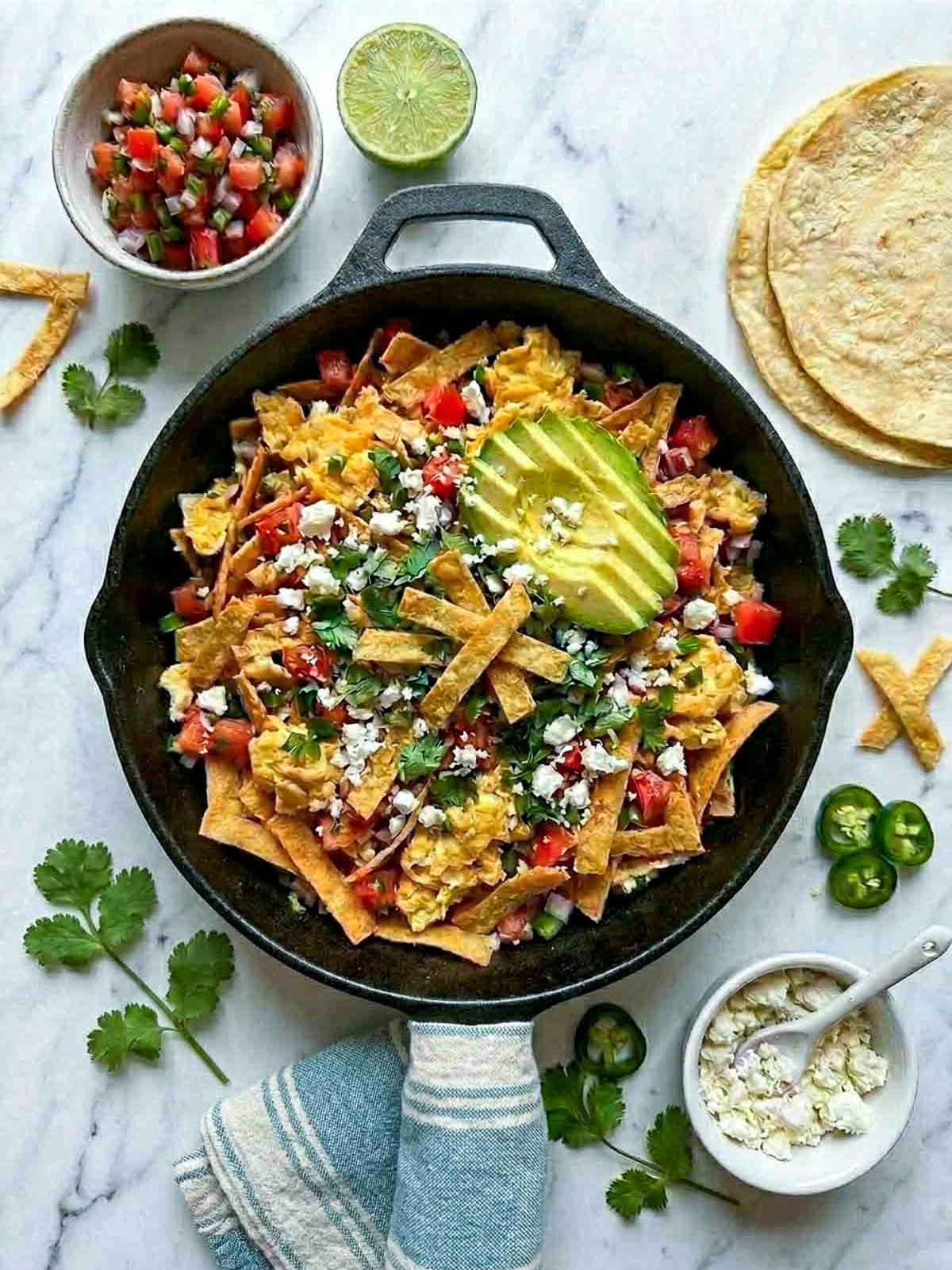 Overhead shot of a cast iron skillet filled with migas — scrambled eggs with crispy tortilla strips, diced tomato, onion, and jalapeño, topped with crumbled cotija, sliced avocado, and fresh cilantro, on a white marble surface with a muted striped linen napkin, bowl of pico de gallo, stack of corn tortillas, cotija, halved lime, and jalapeño slices scattered around.