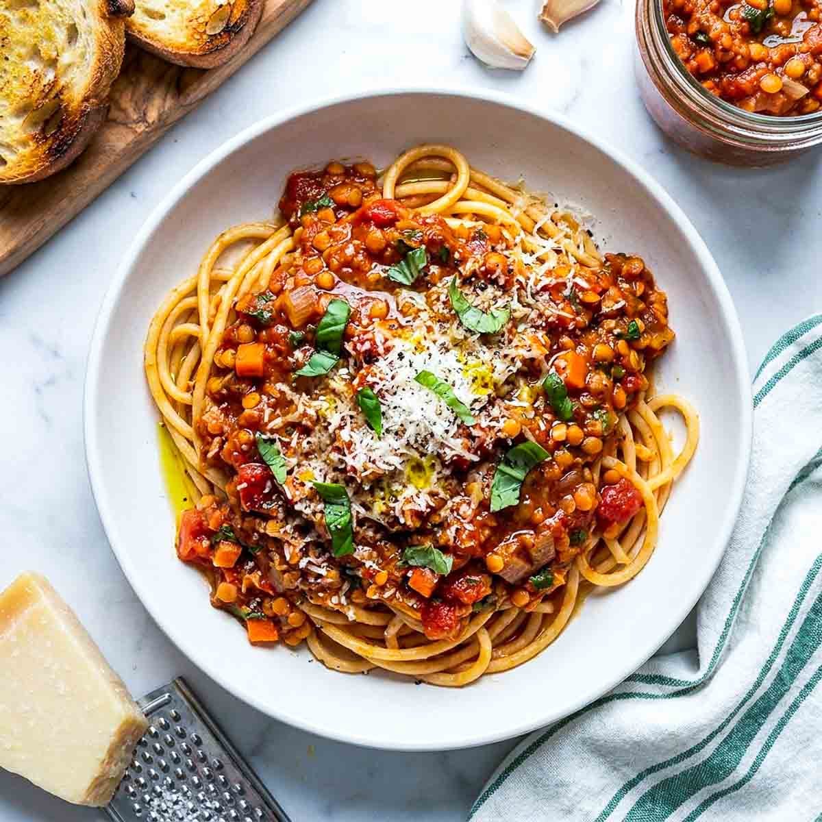 A bowl of spaghetti topped with hearty red lentil Bolognese sauce, grated Parmesan cheese, fresh basil, and a drizzle of olive oil on a white marble surface.