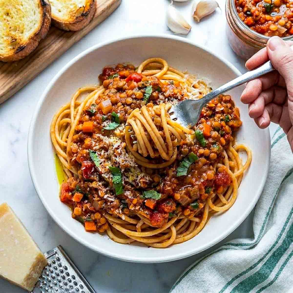 A fork twirling spaghetti coated in rich red lentil Bolognese sauce with Parmesan cheese and fresh basil garnish.