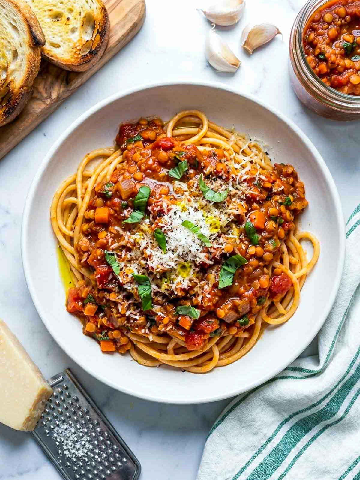 Top-down view of spaghetti served with red lentil Bolognese sauce, Parmesan cheese, fresh basil, toasted bread, and extra sauce on the side.
