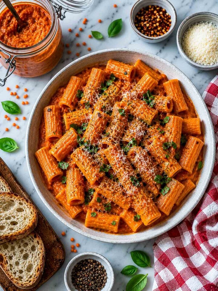 Top-down view of creamy red lentil rigatoni pasta garnished with Parmesan, basil, and black pepper with toasted bread, sauce jar, and red check napkin nearby.