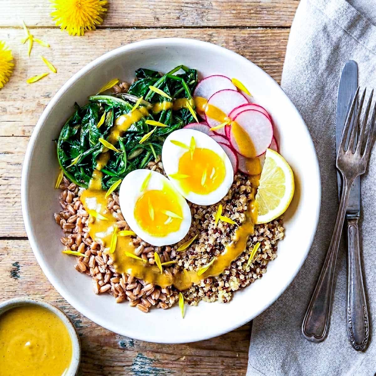
Overhead view of a spring grain bowl with farro, quinoa, wilted dandelion greens, sliced radishes, jammy soft boiled egg halves, lemon wedge, and a golden turmeric tahini drizzle, a small bowl of tahini sauce and dandelion flowers beside it, vintage cutlery on a linen napkin, warm spring light