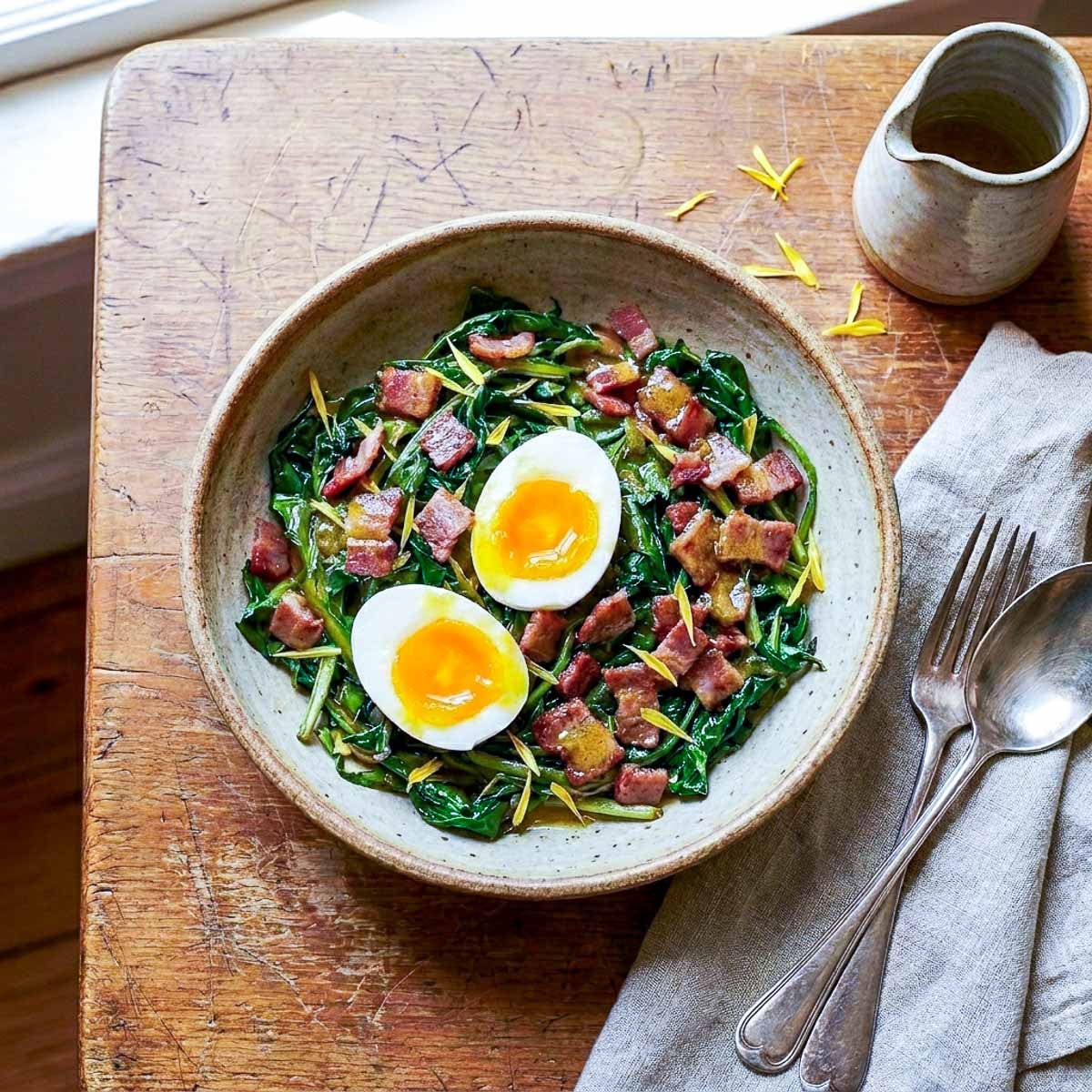 Overhead view of a rustic ceramic bowl of wilted dandelion greens with crispy bacon lardons, two jammy soft boiled egg halves, and scattered dandelion petals, a small dressing jug and vintage cutlery on a linen napkin, warm window light