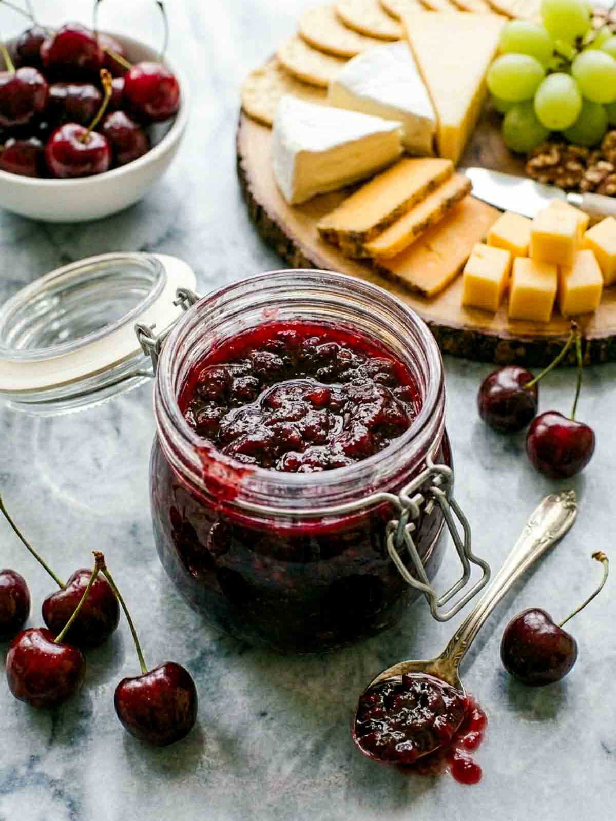 Jar of homemade cherry chutney on a marble surface with fresh cherries, a spoonful of chutney, and a cheese board with crackers, grapes, and assorted cheeses.