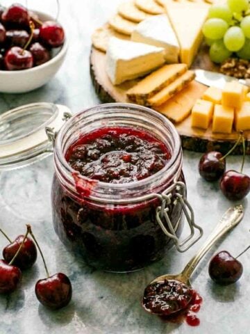 Homemade cherry chutney in an open glass jar served alongside a cheese board with brie, cheddar, crackers, grapes, and fresh cherries.