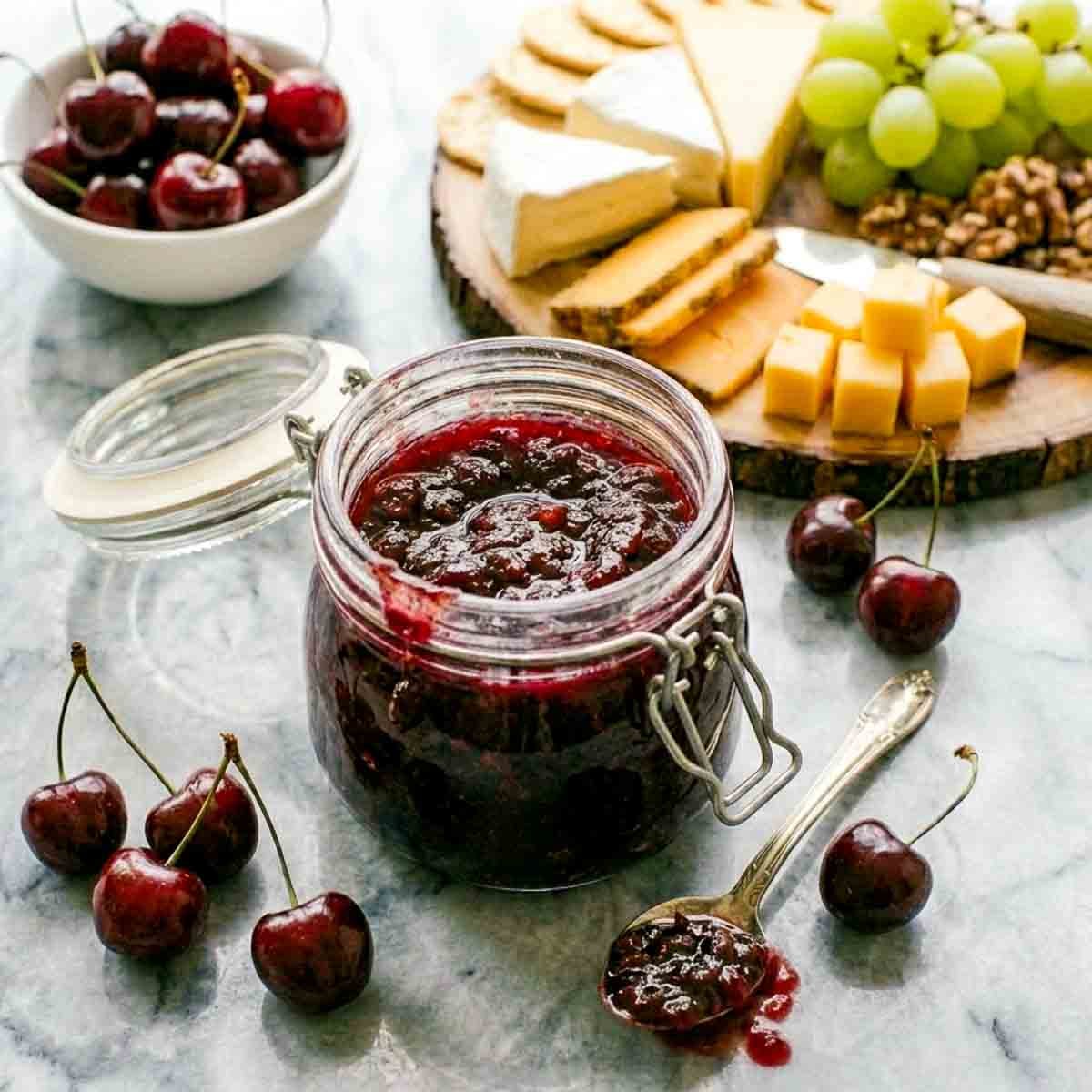 Homemade cherry chutney in an open glass jar served alongside a cheese board with brie, cheddar, crackers, grapes, and fresh cherries.