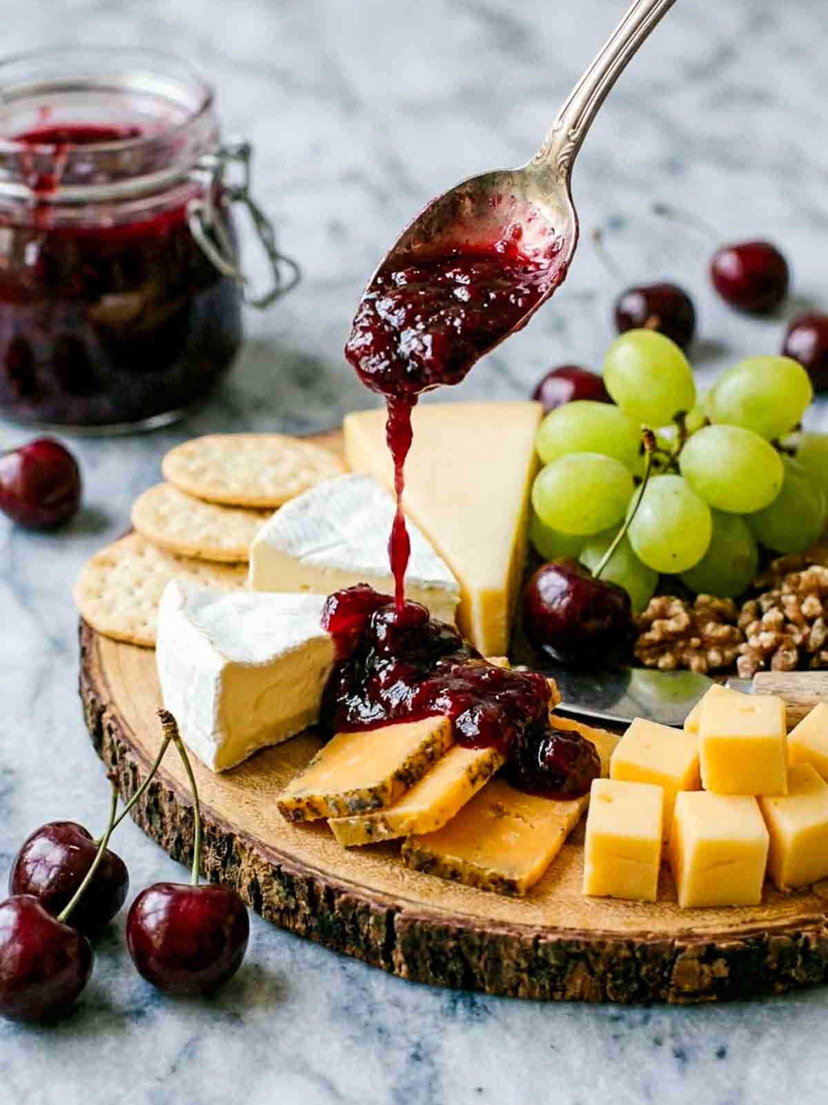 A spoon drizzling homemade cherry chutney over a cheese board with brie, cheddar, crackers, grapes, walnuts, and fresh cherries.
