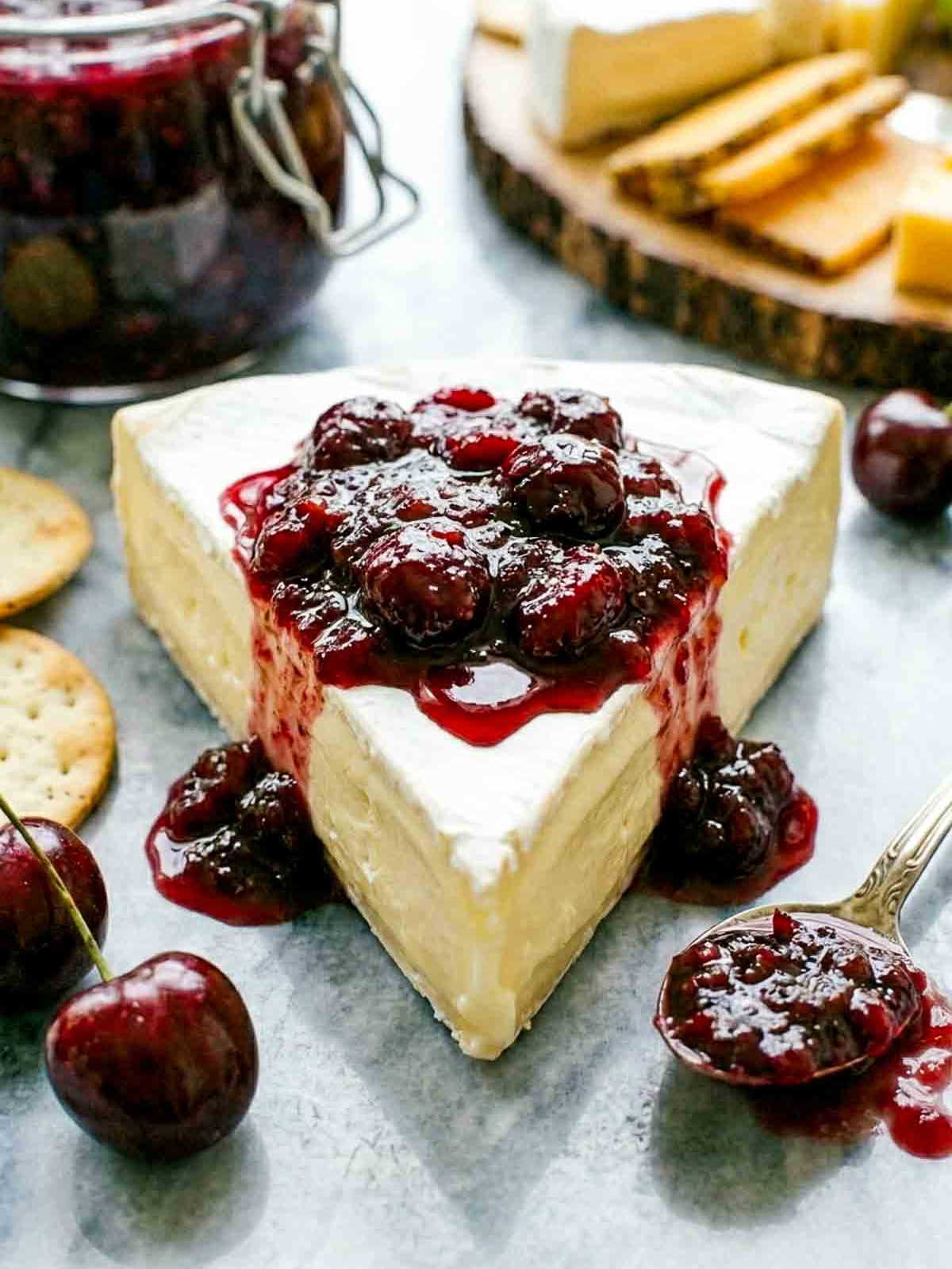 Creamy brie topped with homemade cherry chutney on a marble surface with fresh cherries, crackers, and a jar of chutney in the background.