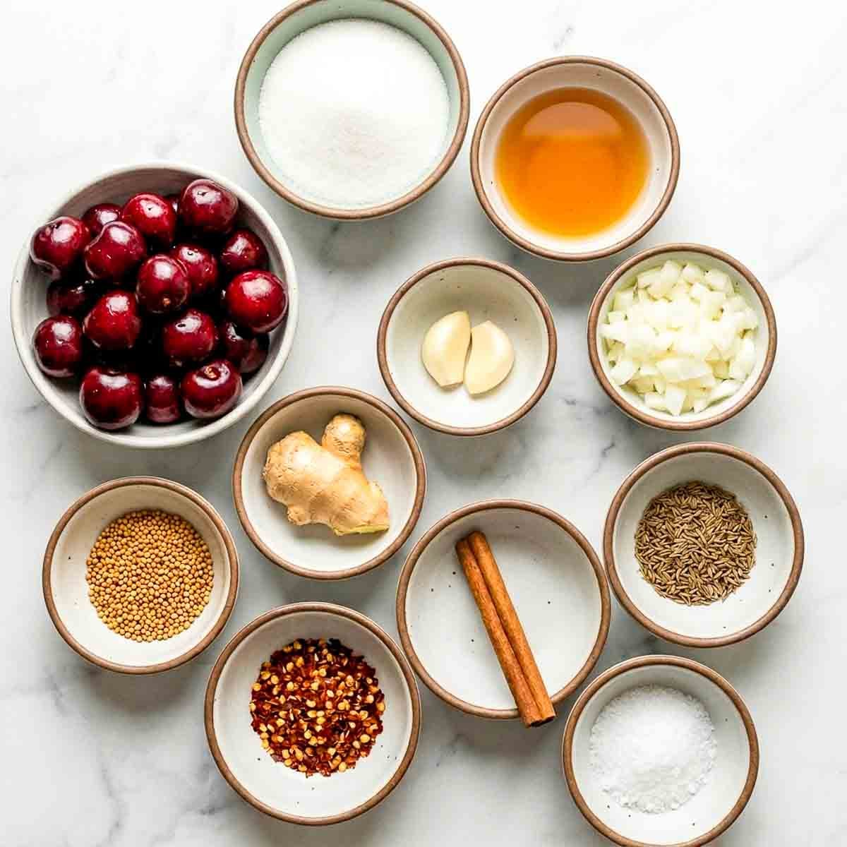 Ingredients for cherry chutney arranged in small bowls, including fresh cherries, onion, garlic, ginger, vinegar, sugar, spices, and salt.
