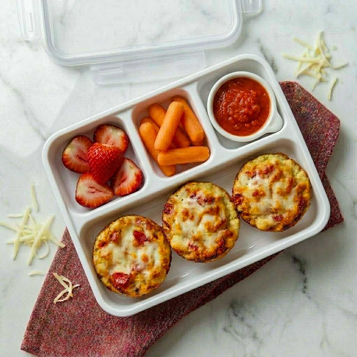 Lunchbox pizza muffins packed in a divided lunch container with marinara dipping sauce, carrot sticks, and strawberries.