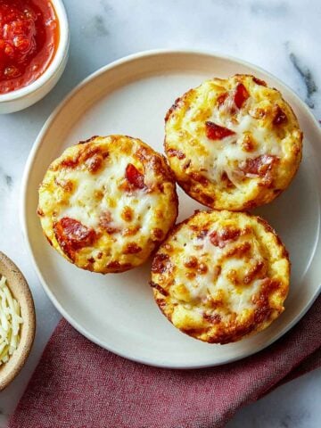 Three cheesy lunchbox pizza muffins topped with melted mozzarella and pepperoni pieces served on a plate with marinara sauce.