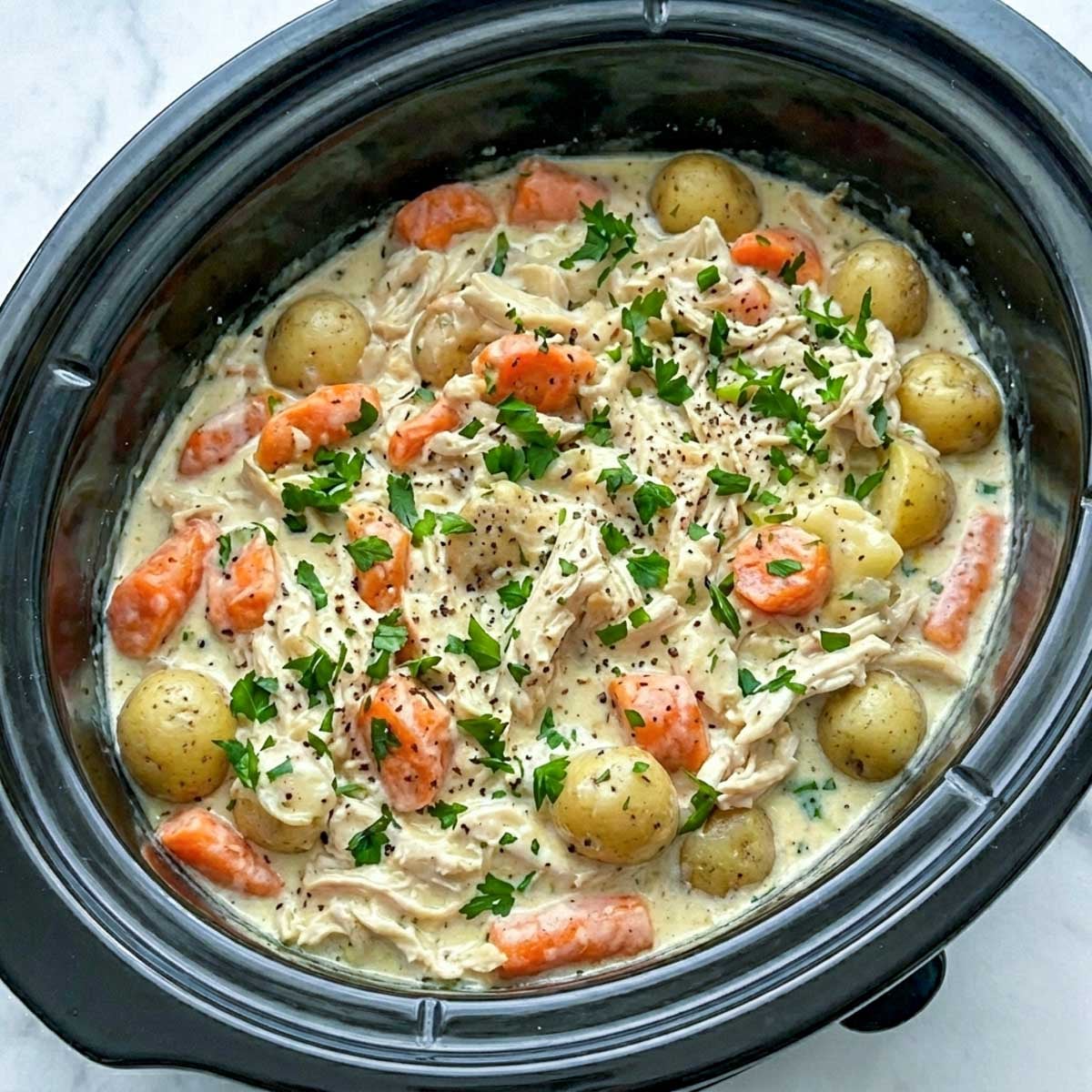 Finished crockpot ranch chicken and potatoes with carrots in a thick creamy ranch sauce, garnished with fresh parsley and black pepper directly in the slow cooker.